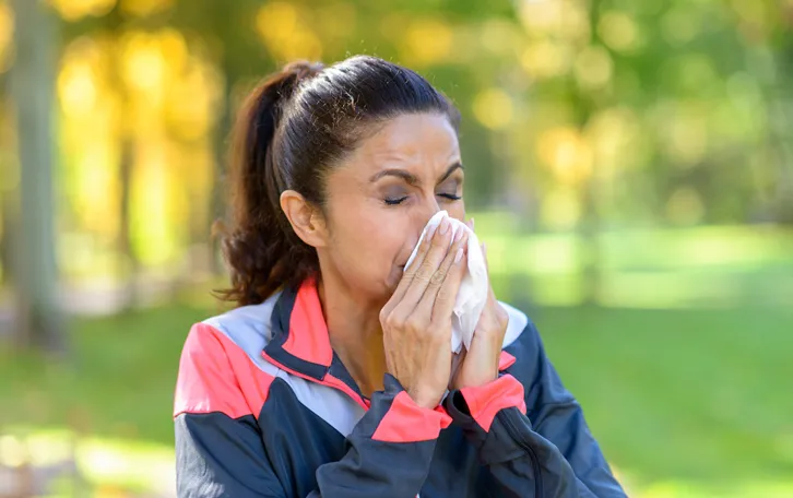 Eine Frau mit geschlossenen Augen in einem dunklem Trainingsanzug putzt sich in der Natur die Nase