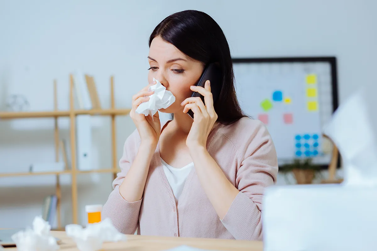 Frau am Telefon mit Taschentuch vor der Nase und Medikamenten vor sich